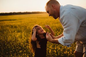 Couple sharing a joyful moment in a golden sunlit flower field at sunset, woman laughing as man leans toward her.