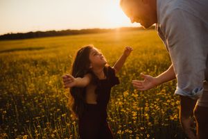 A young girl reaches up with open arms toward an adult in a golden sunlit flower field at sunset.