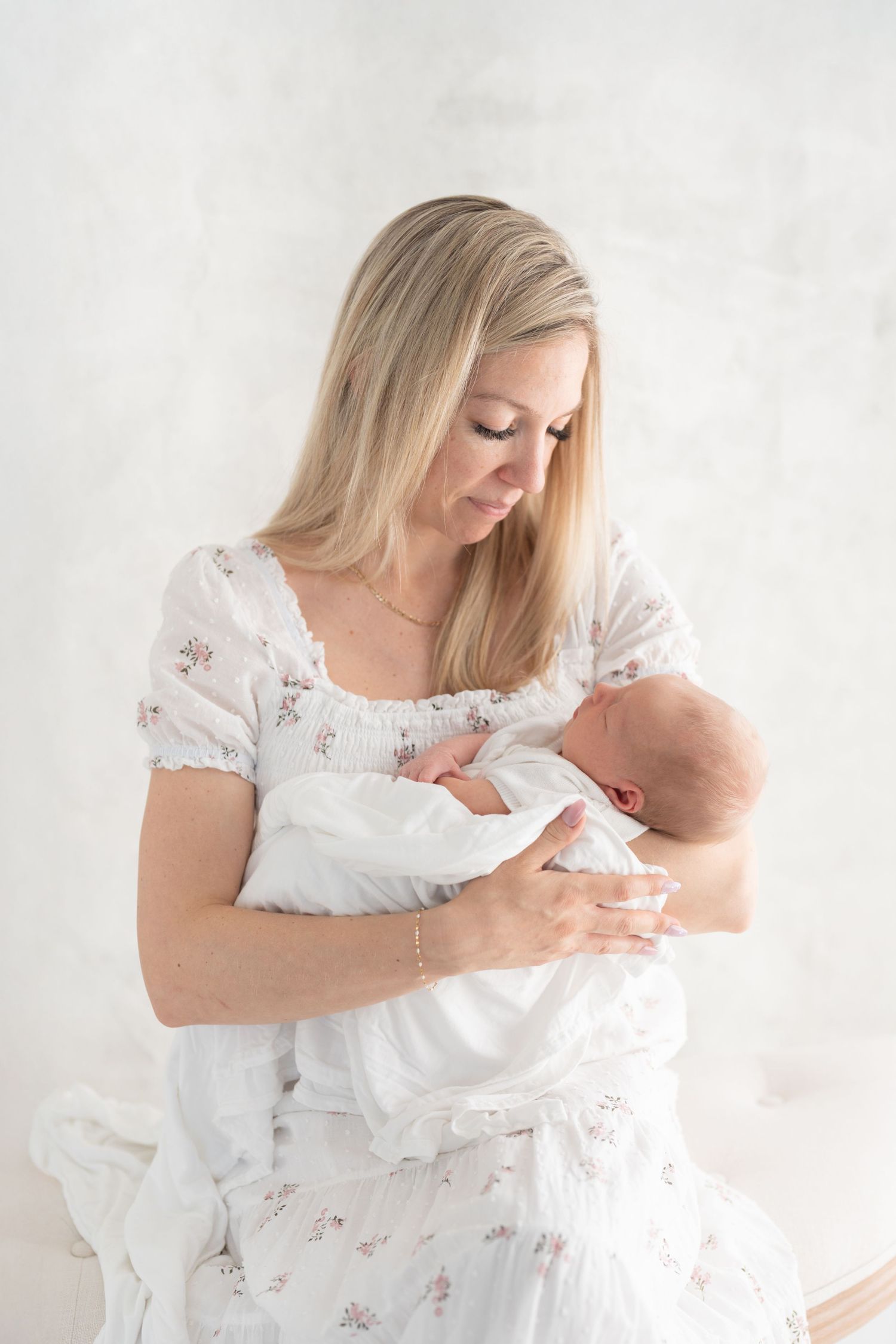 Nouvelle maman tenant son nouveau-né dans une robe blanche et fleurie pour une séance maternité fine art au studio de Québec (St-Augustin)