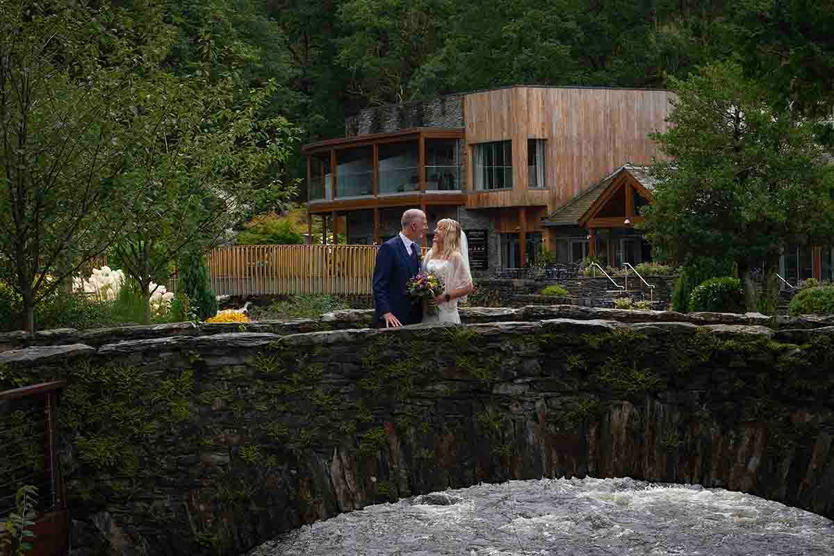 Bride and groom share a romantic moment on a stone bridge surrounded by lush greenery and a rustic wooden venue.