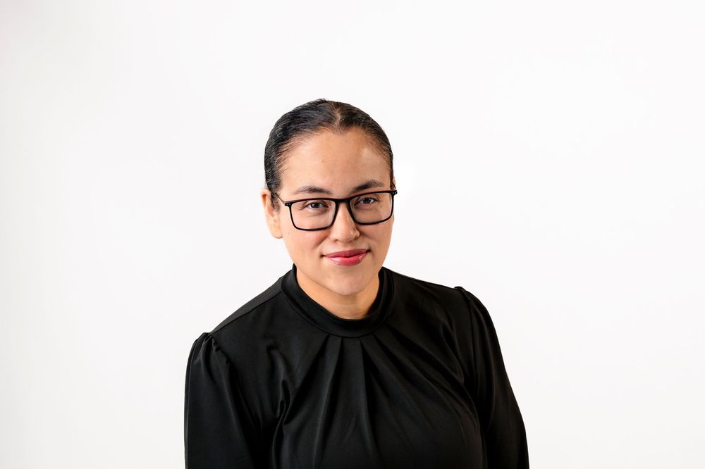 Woman with glasses wearing a black top, smiling against a white background.