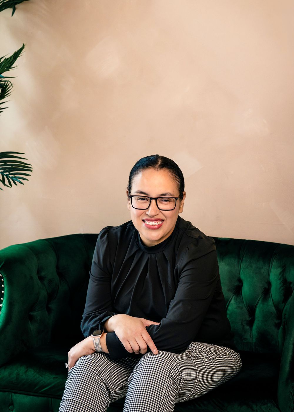 Smiling woman with glasses in black top and houndstooth pants sitting on a green velvet tufted sofa.