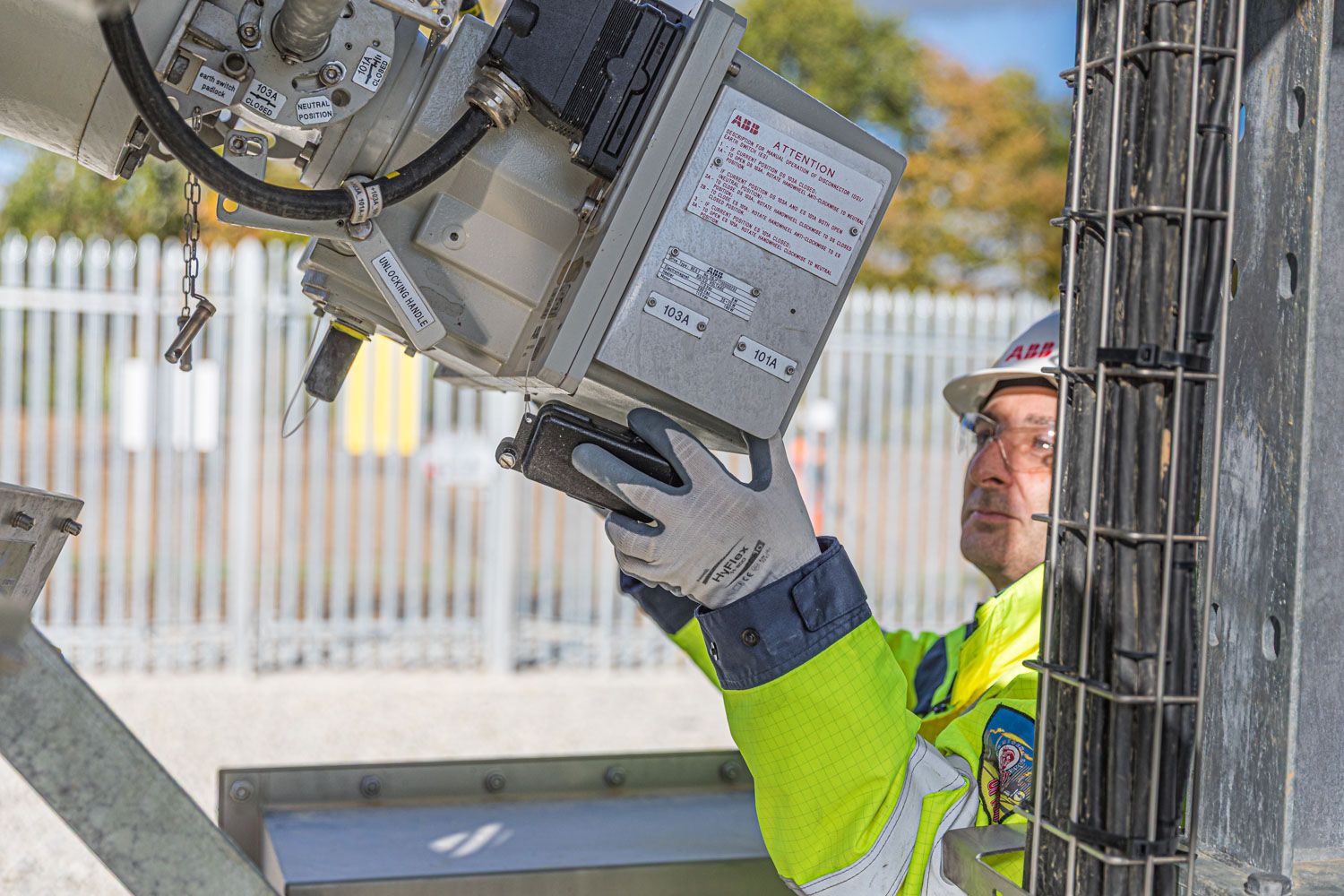 A worker in high-visibility gear installs power equipment on a metal pole at an outdoor facility.