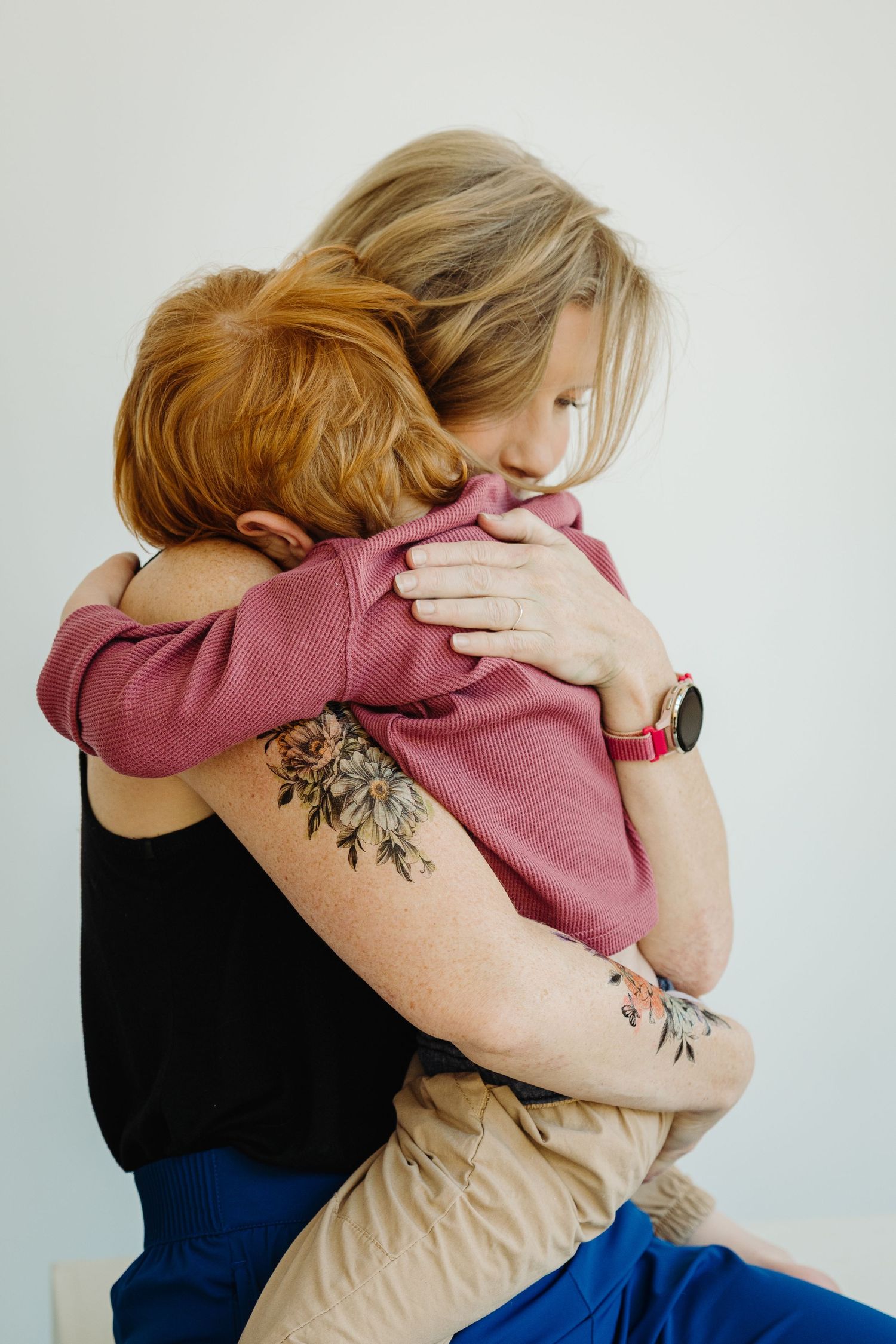 A tattooed woman with a watch hugs a child in a pink shirt against a white background.