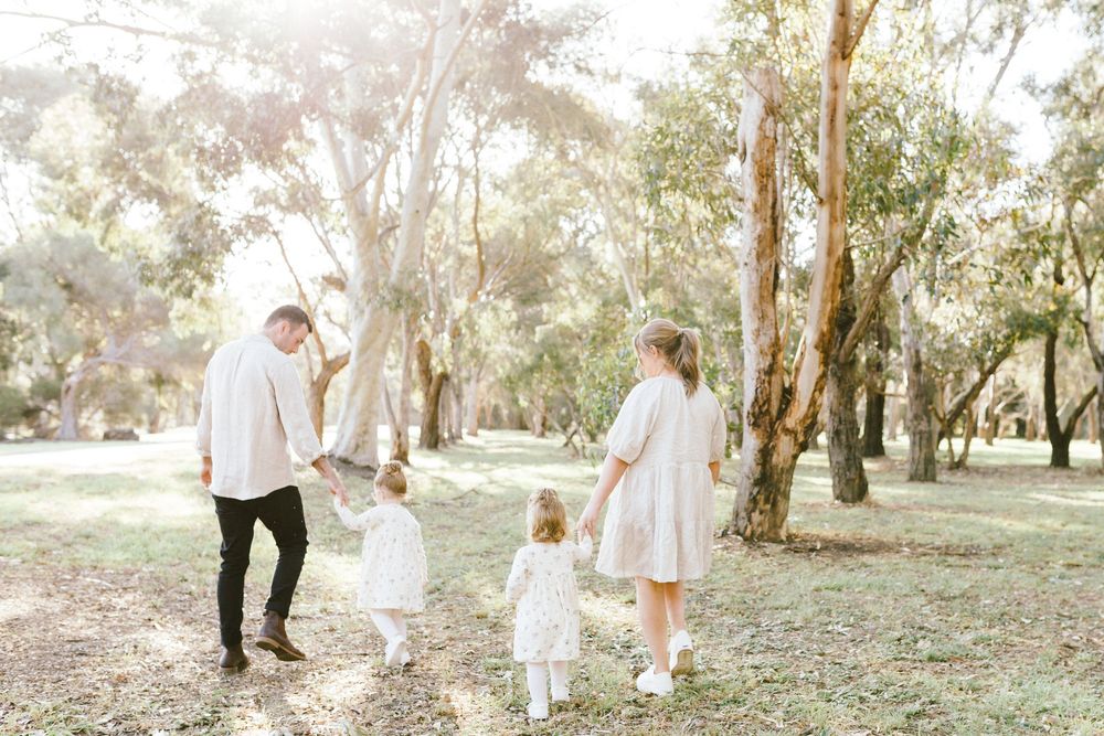 Family of four walking together in a sunlit park with tall eucalyptus trees, dressed in white and neutral tones.