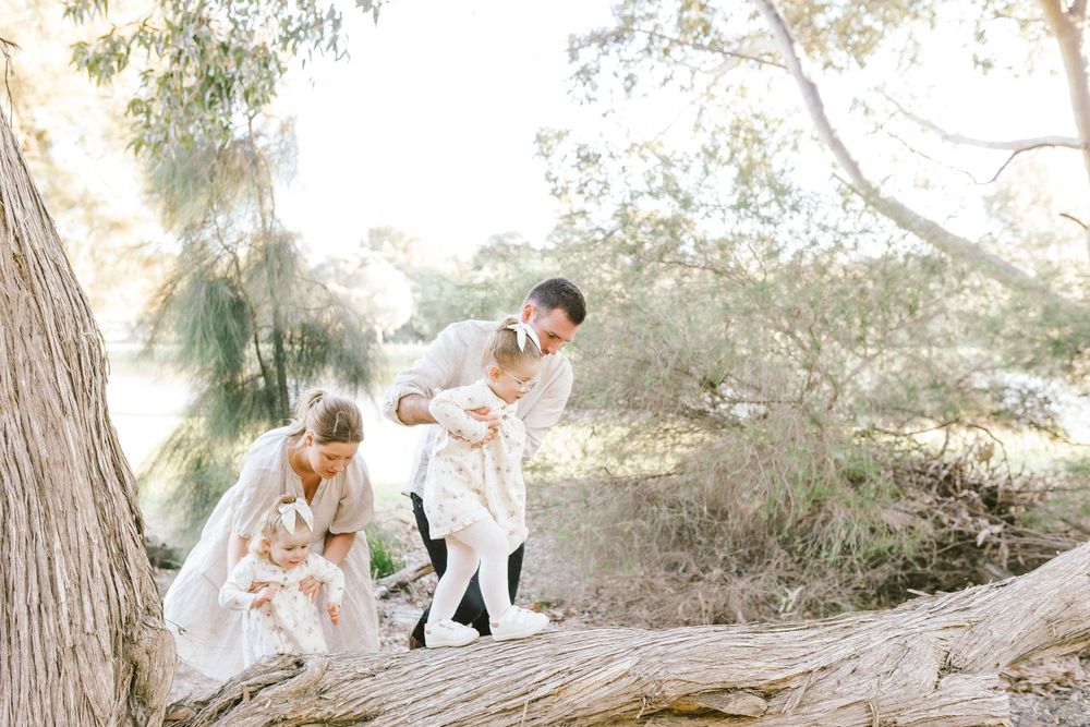 Family in white outfits playing on a fallen log in a sunlit park with lush green trees in the background.