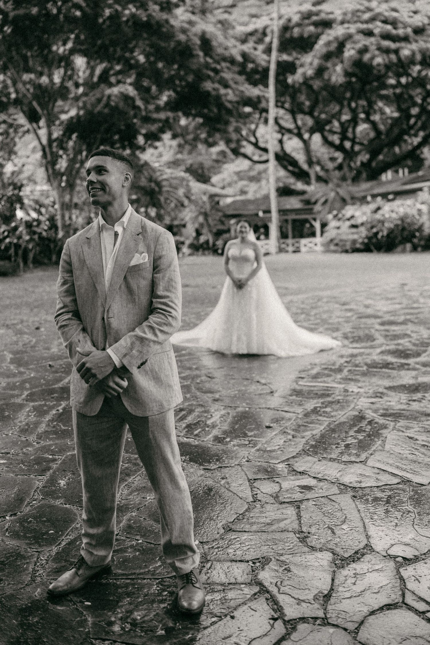Black and white photo of a groom smiling as his bride in a wedding gown approaches across a stone patio.
