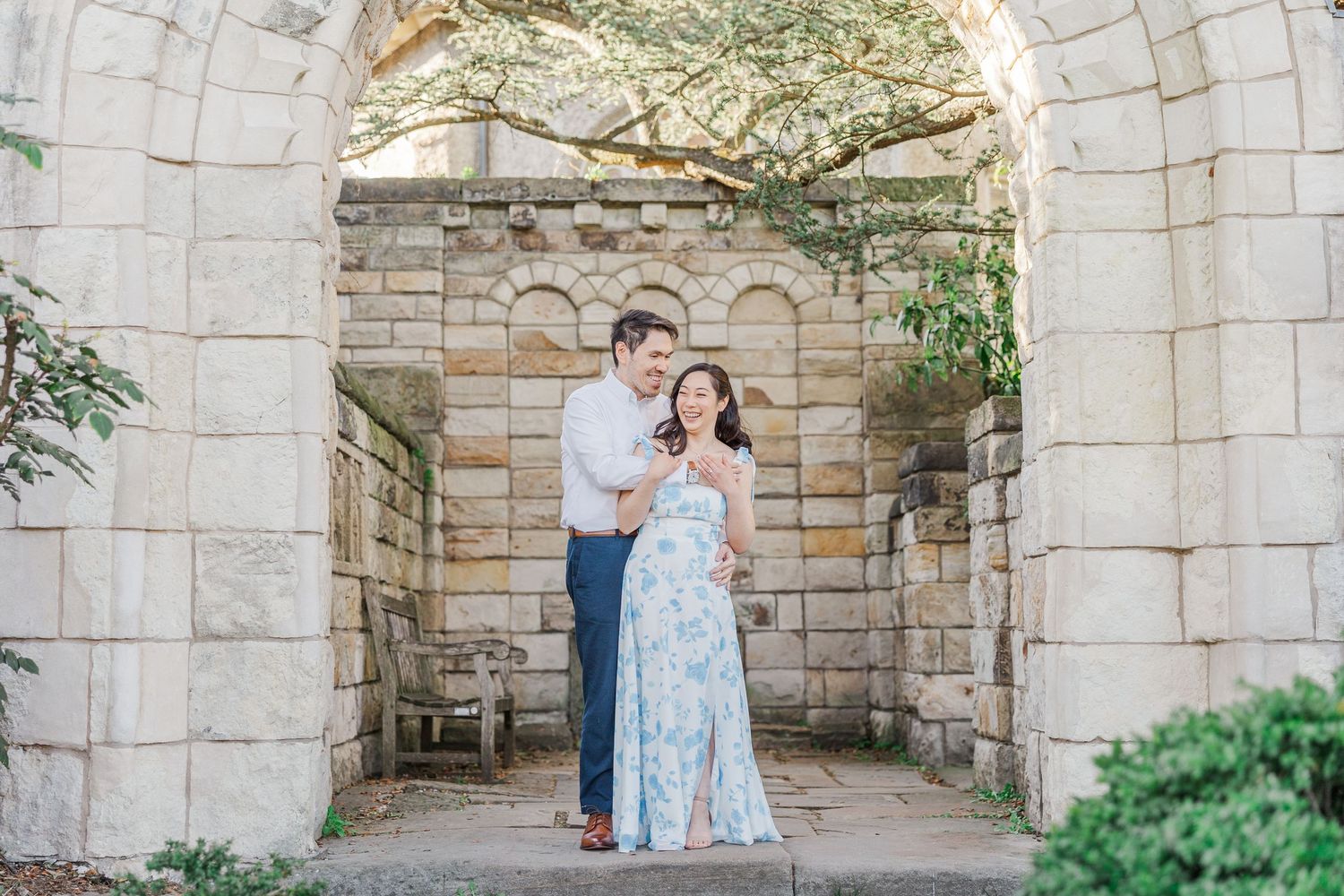Couple posing under stone arch at historic garden venue, woman in floral dress, man in navy pants.