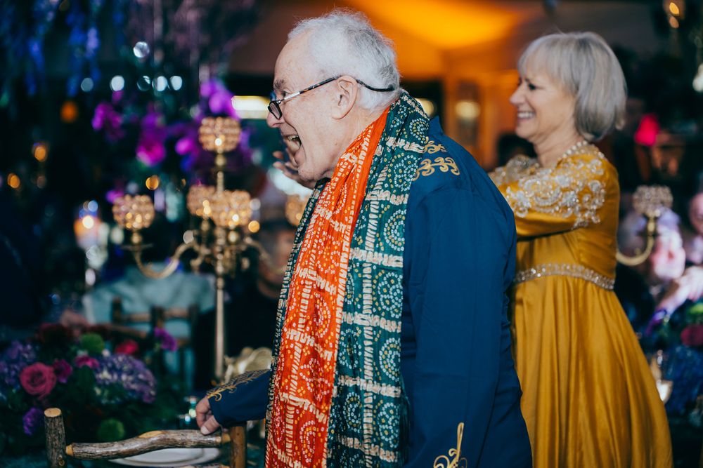 Elderly man in navy suit with colorful orange and green scarf laughing at an elegant Indian-themed event.