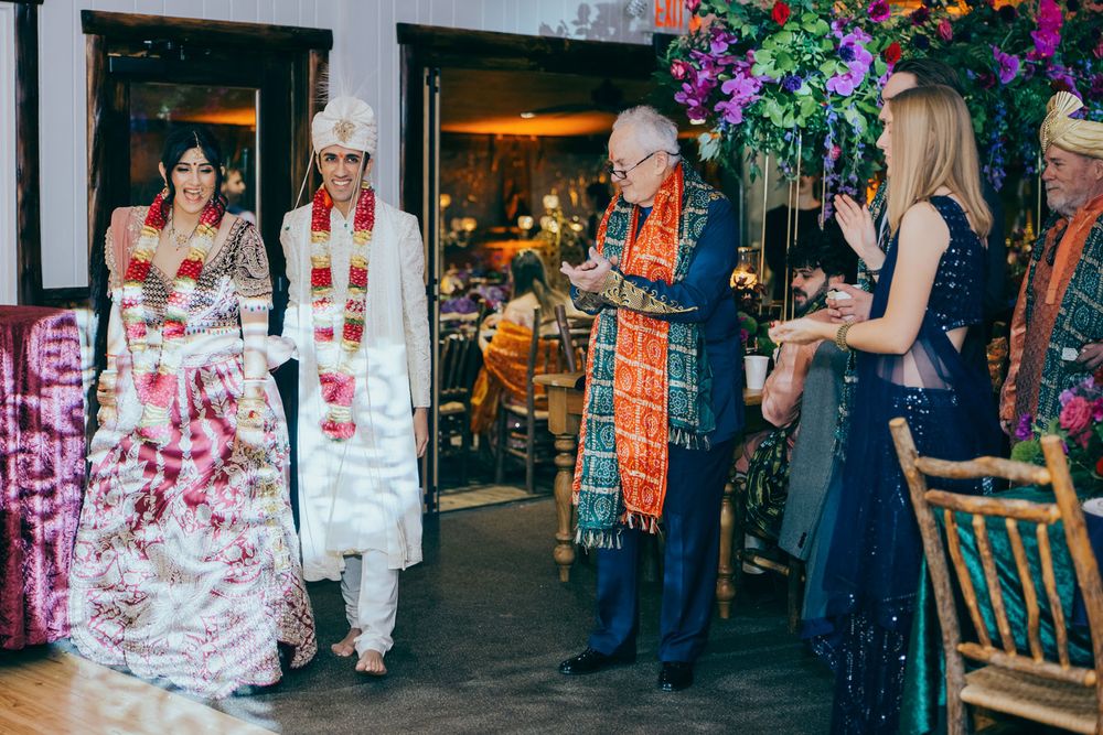 Couple in traditional Indian wedding attire walks down the aisle at a colorful indoor ceremony with guests applauding.