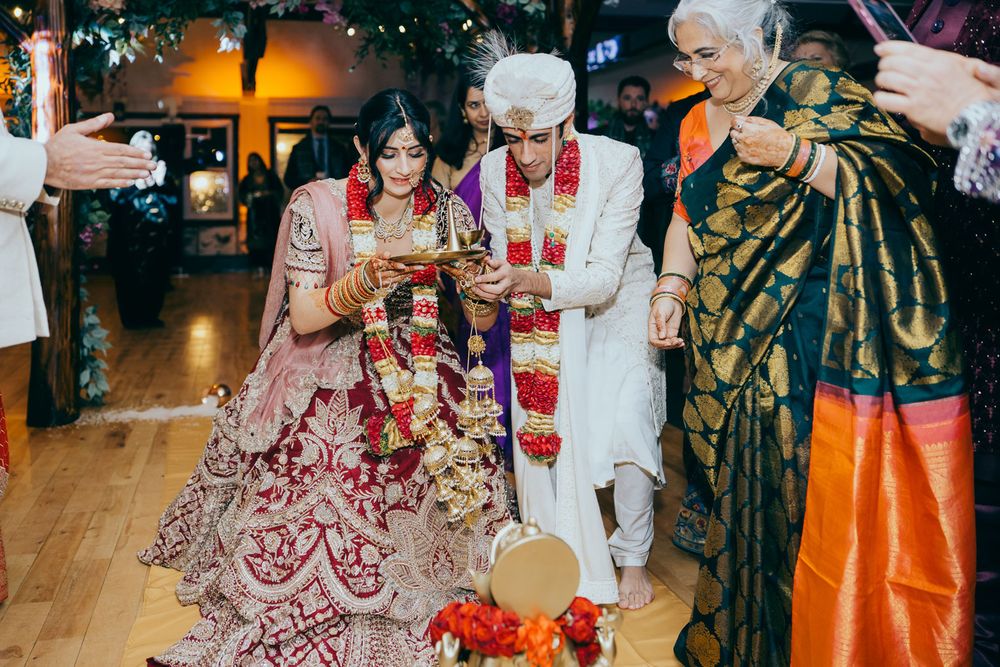 Bride and groom in traditional Indian wedding attire perform ceremony rituals with elder guest on wooden floor.