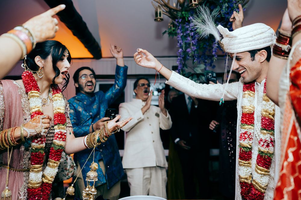 Indian bride and groom in traditional wedding attire exchange garlands joyfully surrounded by guests at a vibrant ceremony.