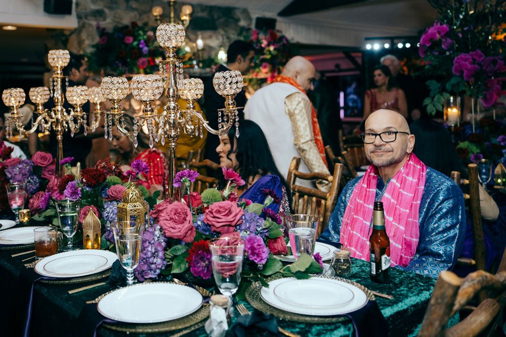 Man in pink jacket seated at elegant floral table with crystal candelabras and colorful roses at formal event.