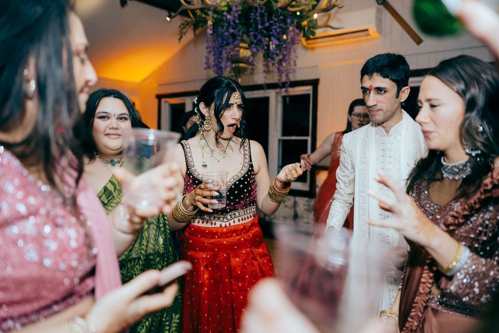Bride in red lehenga and groom in white sherwani celebrate with guests at a vibrant Indian wedding reception.