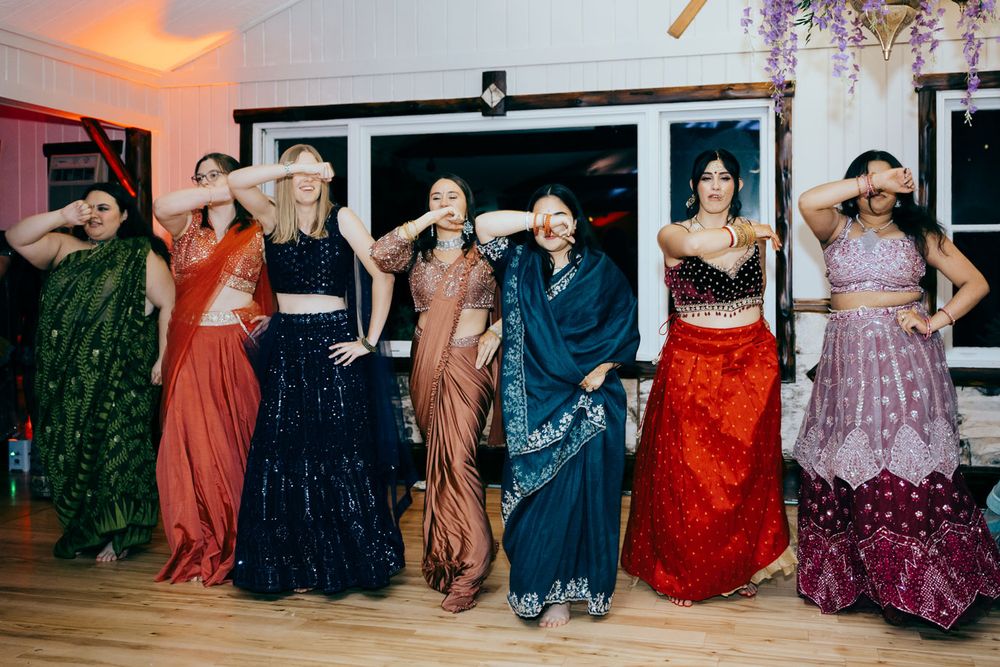 Seven women in colorful Indian attire pose playfully on a dance floor at a festive celebration.