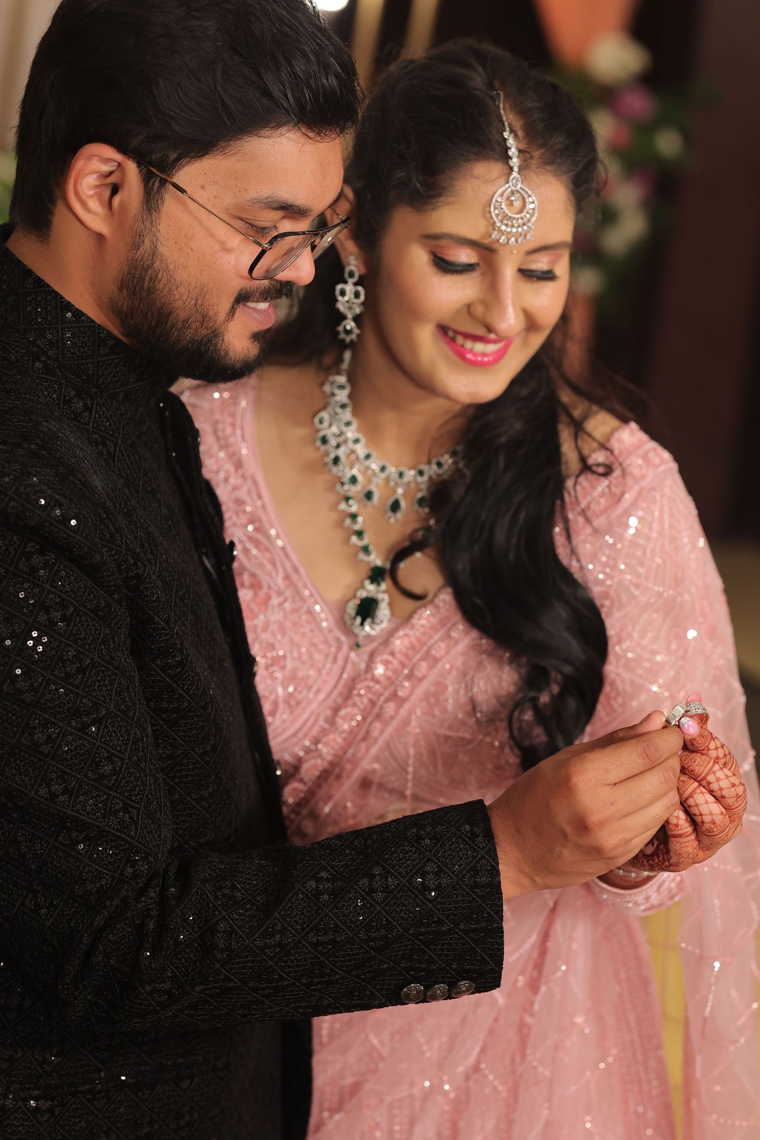 Couple in elegant attire at Indian engagement ceremony, bride in pink saree with diamond jewelry admiring ring.