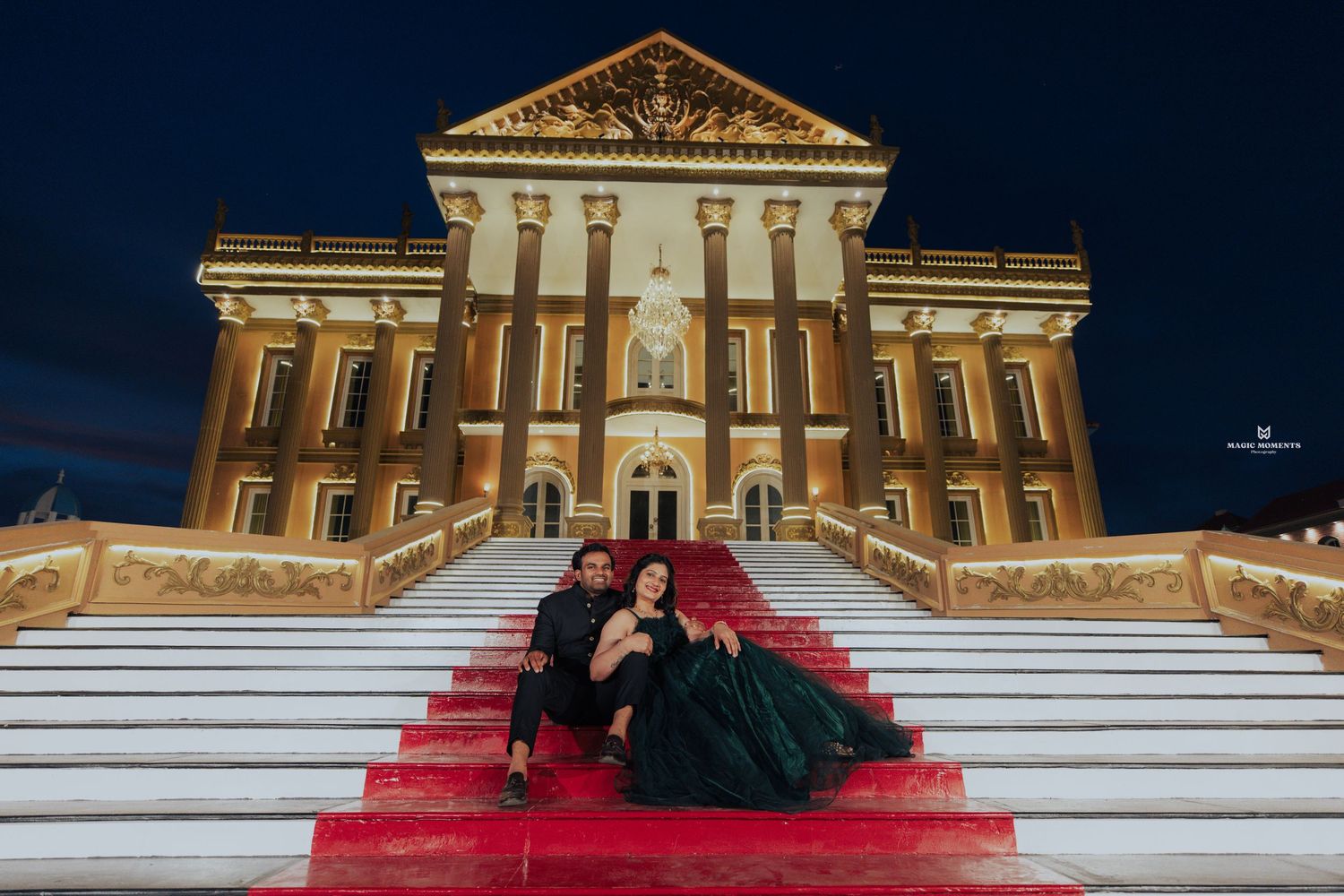 Couple in elegant attire sitting on red-carpeted steps of illuminated classical building at night.