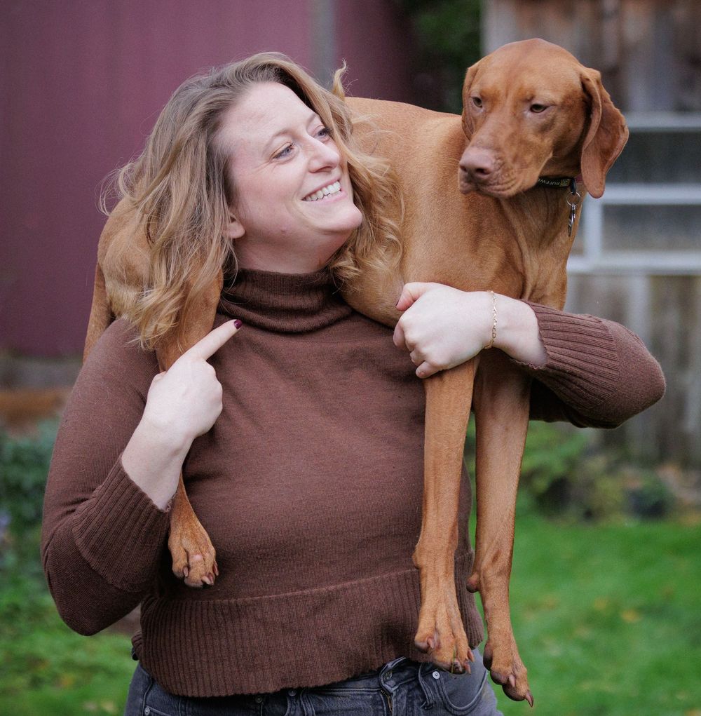 Smiling woman in brown turtleneck carrying a Vizsla dog on her shoulders outdoors.