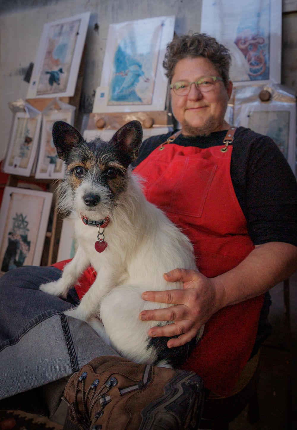 Artist in red apron holding a scruffy Jack Russell terrier dog in a creative studio.