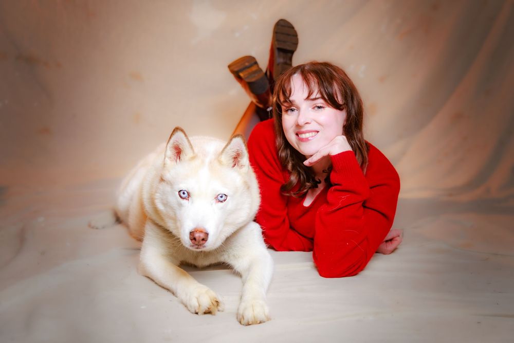 Woman in red sweater with reindeer antlers poses beside a white Siberian Husky with blue eyes on beige backdrop.