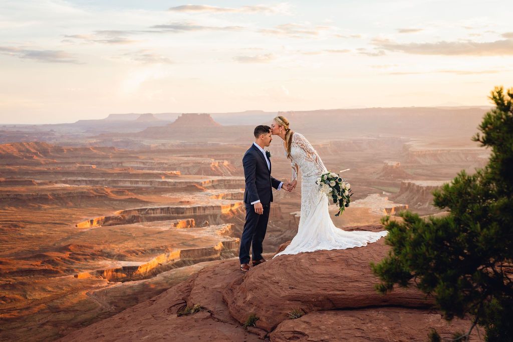 Bride and groom kissing on a red rock cliff overlooking a vast canyon at sunset.