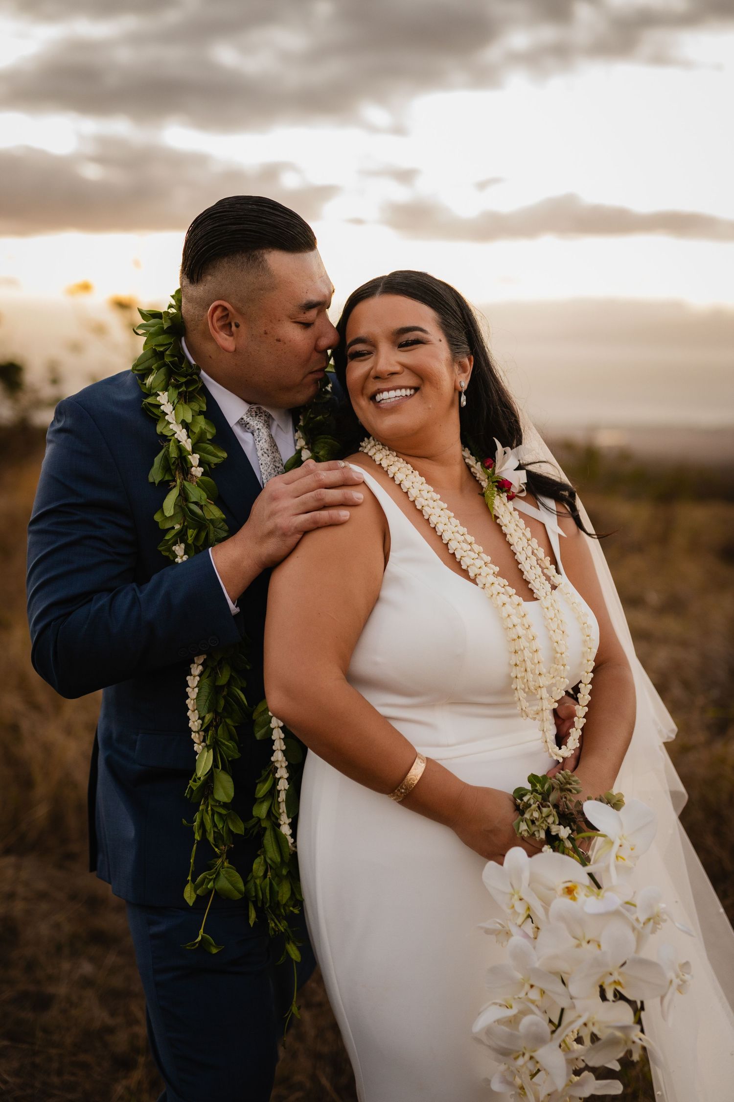 Groom in navy suit kisses smiling bride wearing floral lei at sunset in open field. hawaii wedding photography