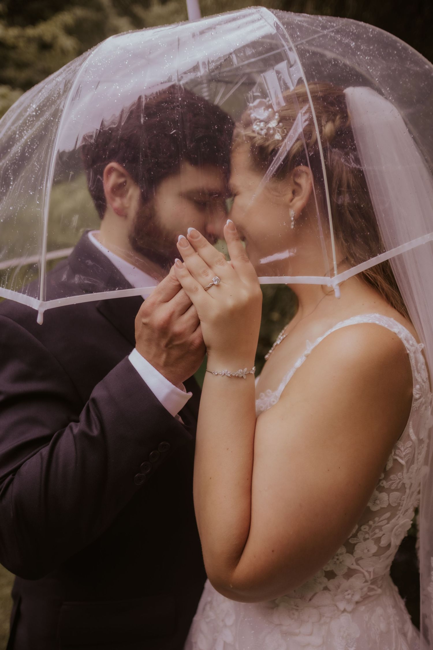 Bride and groom share romantic moment under clear umbrella in rain, showing wedding ring, wearing white lace gown and dark suit.