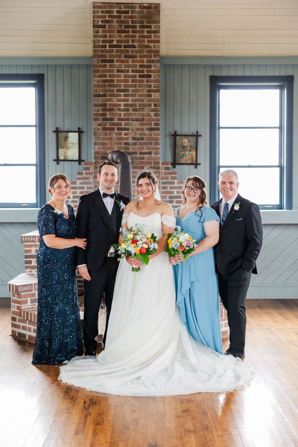 Wedding party poses indoors with bride in white gown holding colorful bouquet, flanked by family in navy and blue attire.