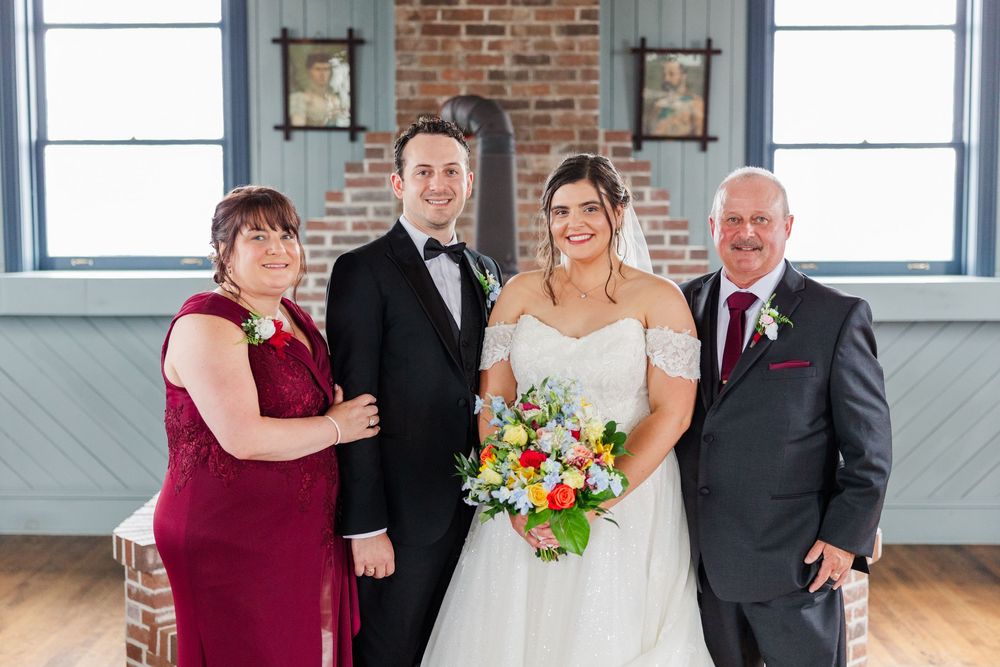 Bride and groom pose with family in elegant wedding attire, bride holding colorful bouquet in a rustic brick venue.
