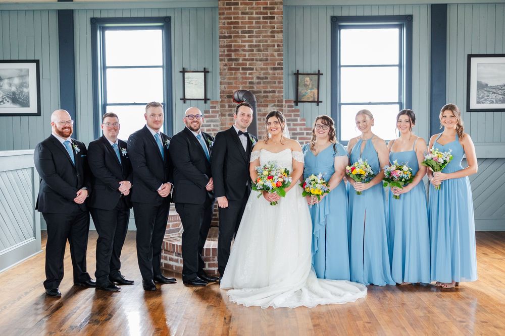 Wedding party poses indoors, groomsmen in black suits, bridesmaids in blue dresses, bride in white gown holding colorful bouquet.