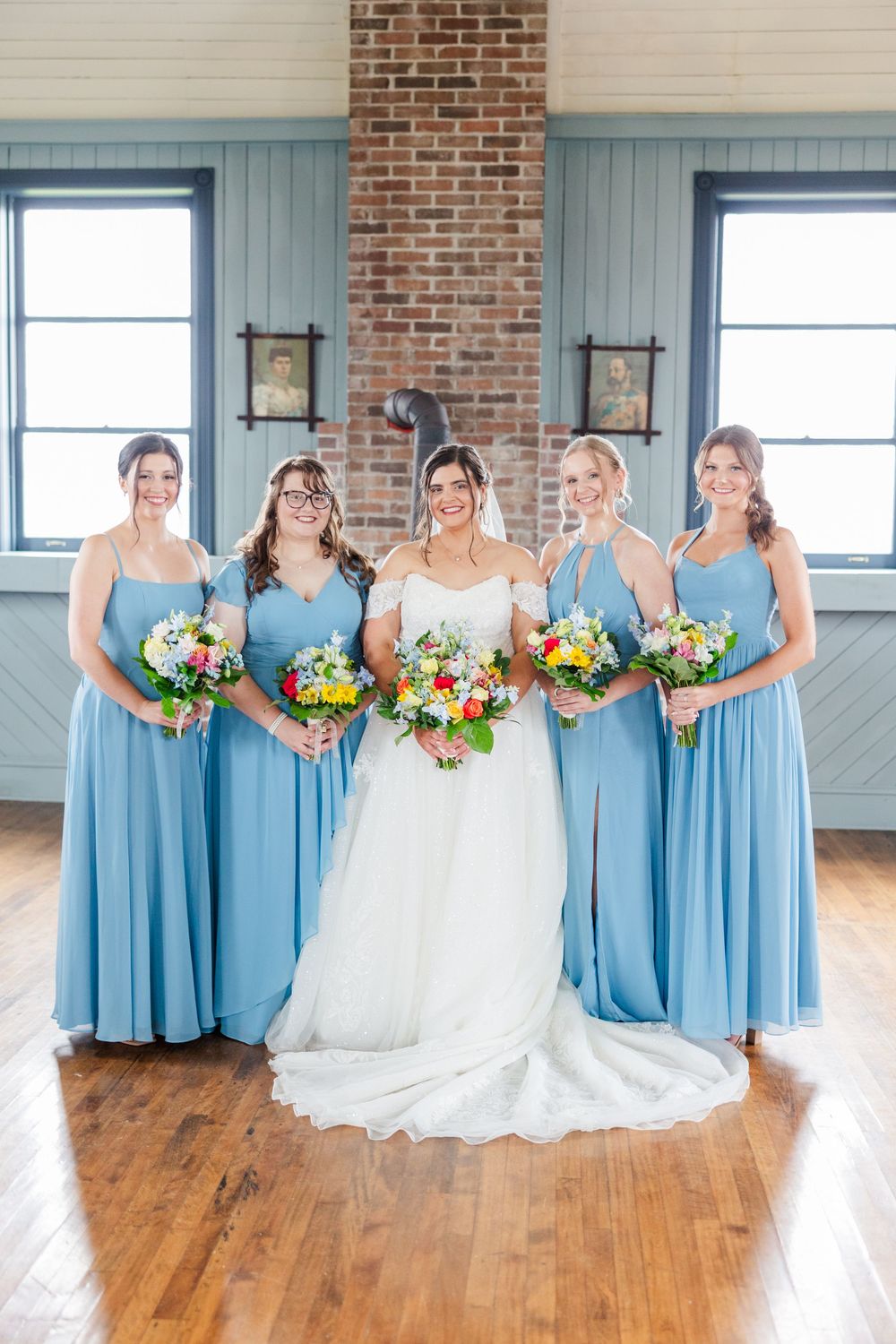 Bride in white gown with four bridesmaids in blue dresses holding colorful bouquets indoors.