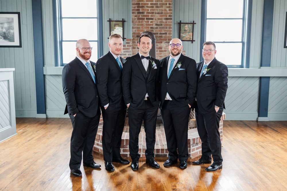 Five groomsmen in black suits with teal boutonnieres pose together smiling in a rustic wedding venue.