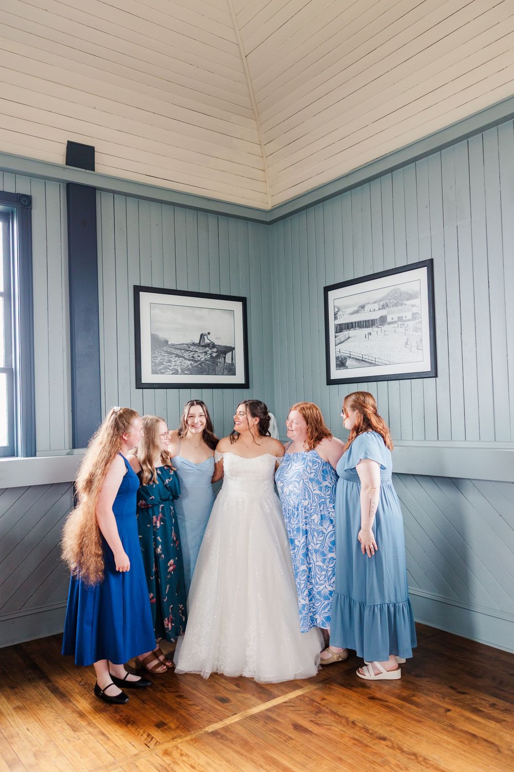 Bride in white gown surrounded by four bridesmaids in blue dresses laughing inside a rustic venue.