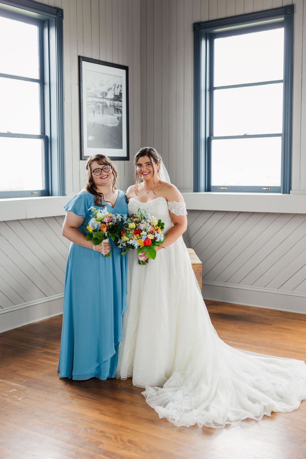 Bride in white gown and bridesmaid in blue dress holding colorful bouquets inside a rustic venue.
