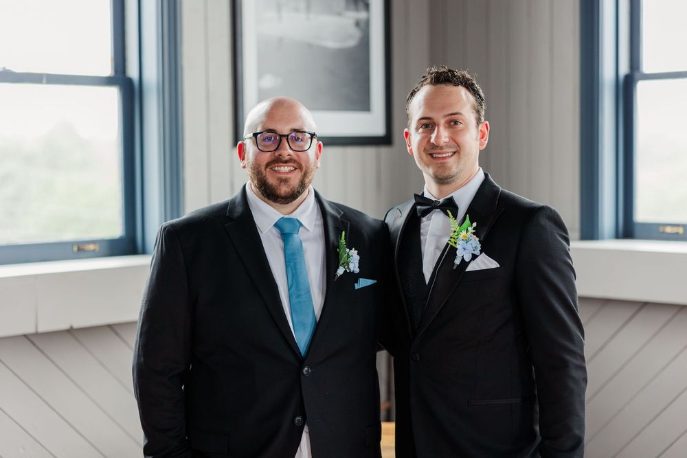 Two grooms in black suits with boutonnieres smiling together at their wedding reception.