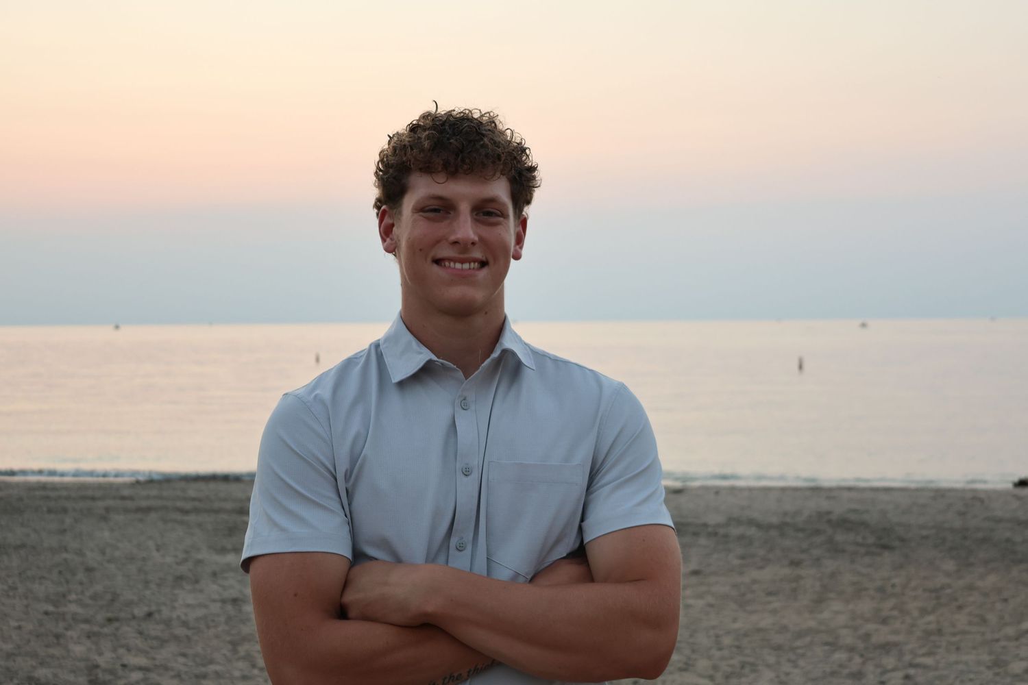 Young man with curly hair smiling on a sandy beach at sunset with arms crossed.