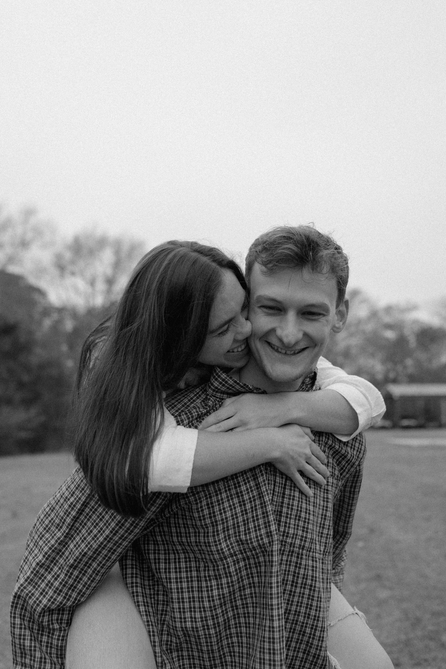 Black and white photo of a smiling man giving a woman a piggyback ride outdoors, sharing a joyful romantic moment.