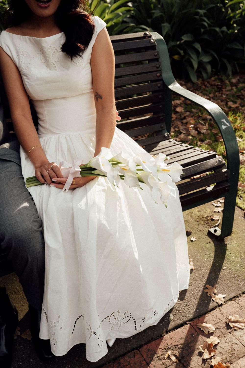 Bride in white lace wedding dress holding calla lily bouquet, seated on garden bench with groom.