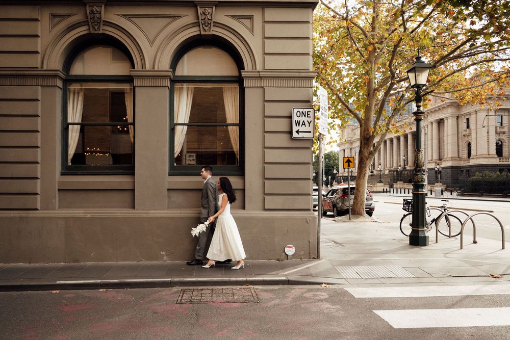 A bride and groom walk hand in hand across a city street corner past a classic stone building.