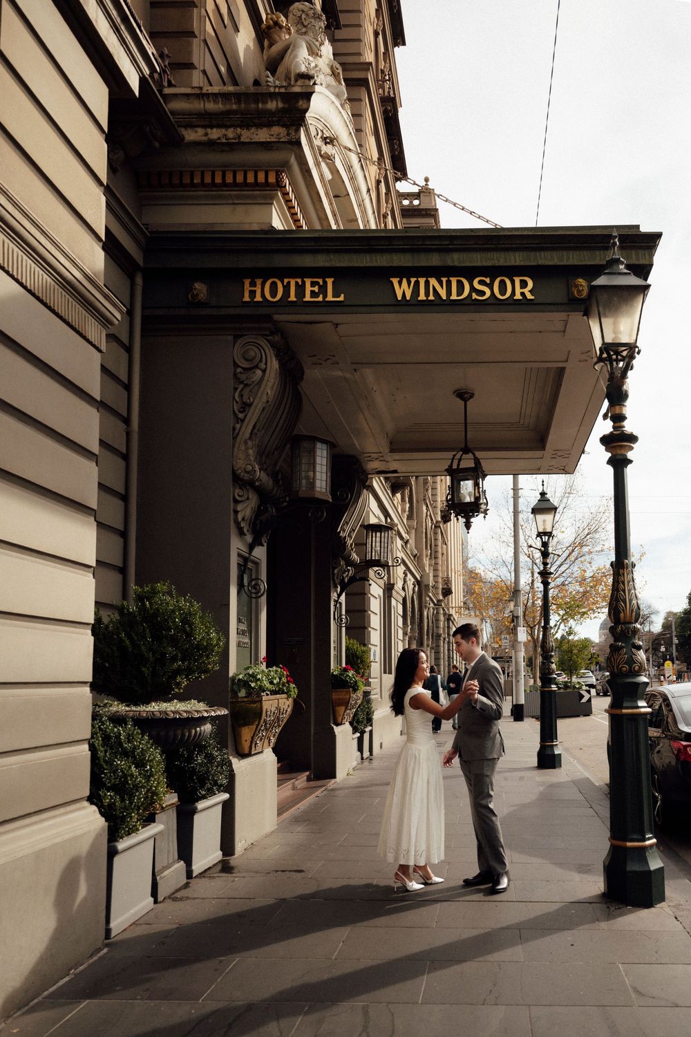 Couple in wedding attire standing outside the historic Hotel Windsor on a sunny day.