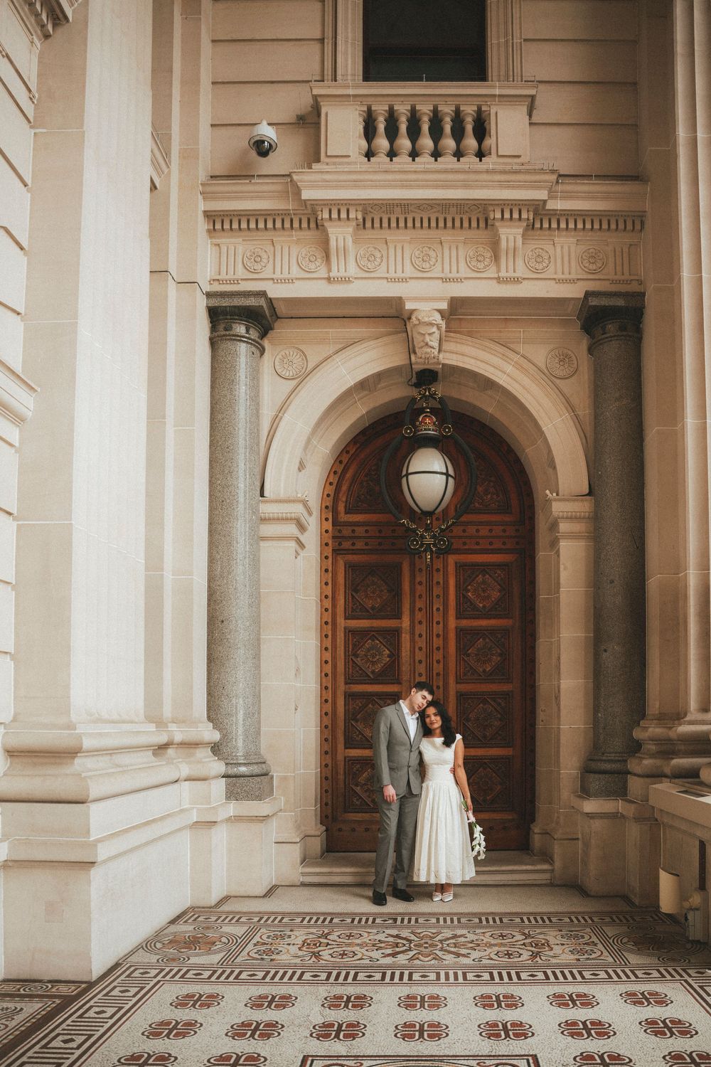 Bride and groom posing in front of ornate wooden door of historic building with elegant columns.
