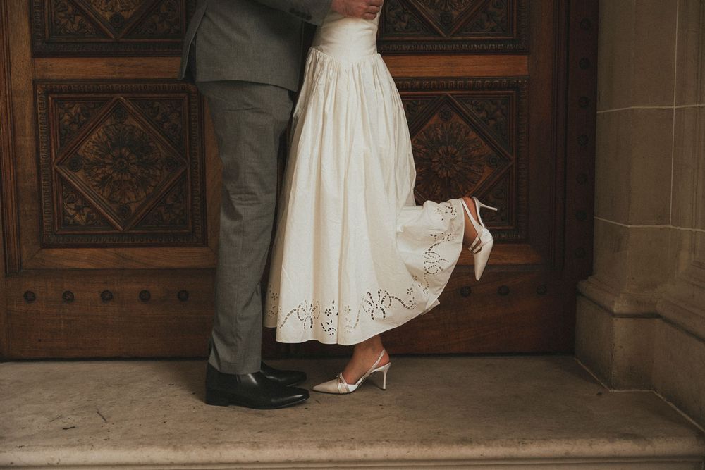 Bride in white lace midi skirt and heels stands with groom in gray suit before ornate wooden church doors.