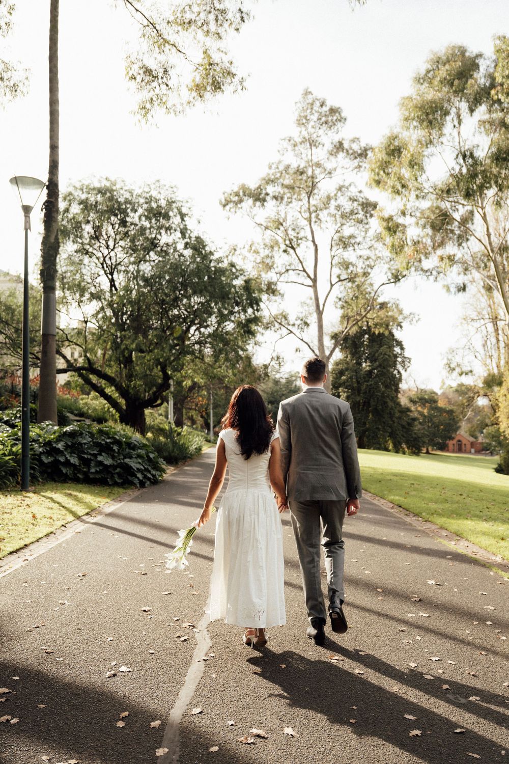 Bride and groom walking hand in hand down a tree-lined park path, bride in white dress holding bouquet.
