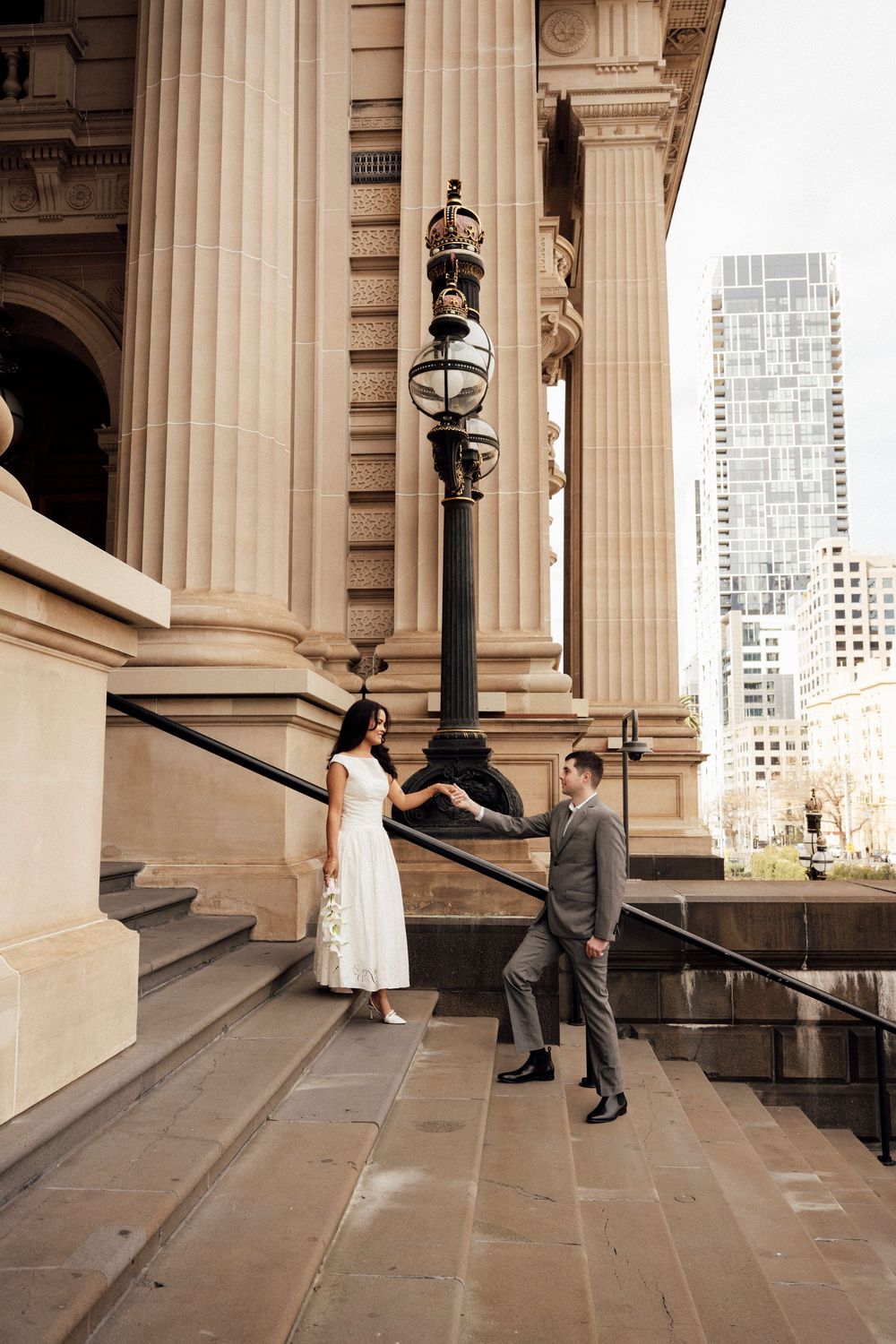 Couple in wedding attire holding hands on steps of historic stone building with ornate lamp post and city skyline.