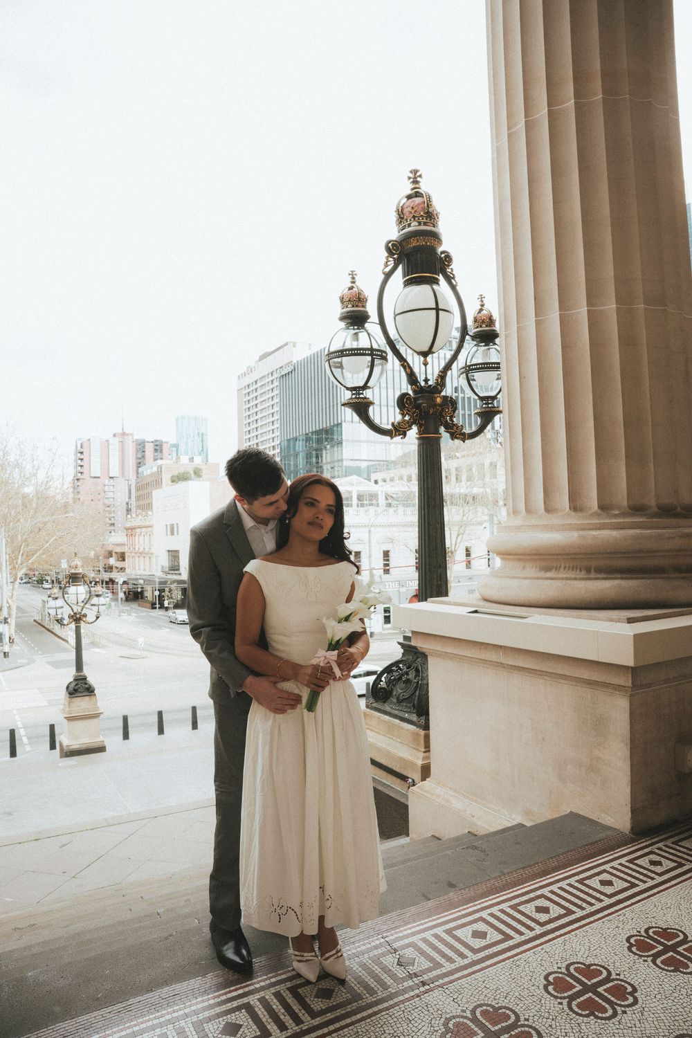 Couple in wedding attire embracing on ornate steps near a decorative lamppost with city buildings in the background.