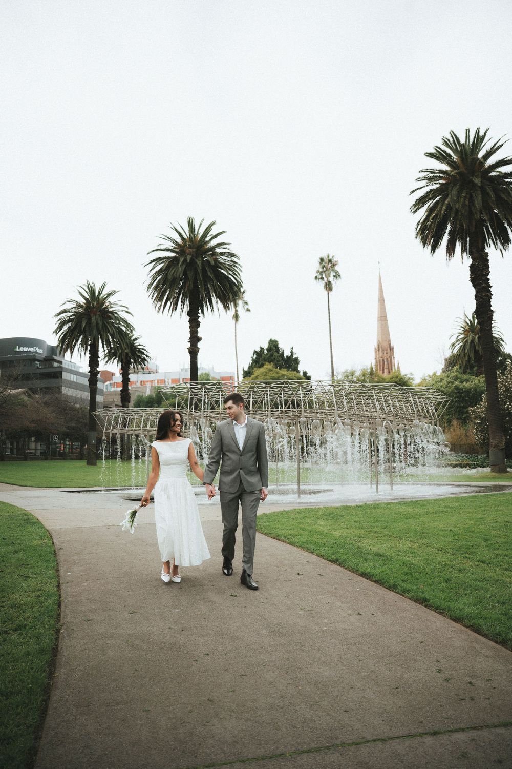 Bride and groom walking hand in hand through a park with palm trees and a fountain backdrop.