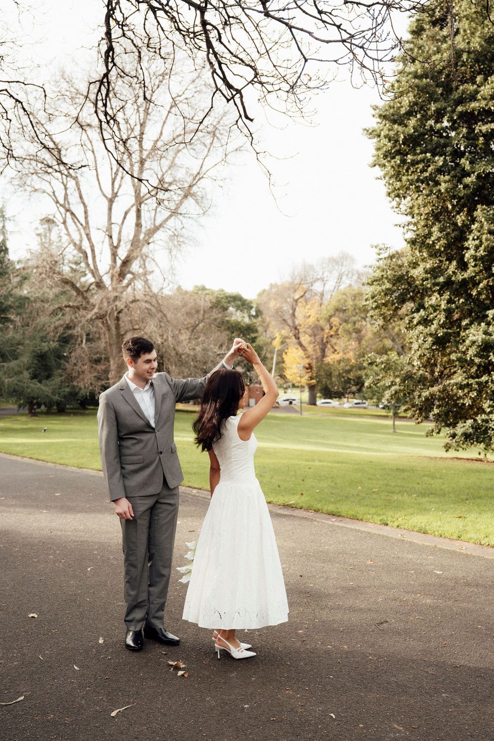 Bride and groom dancing together in a lush green park, bride wearing a white midi dress and heels.