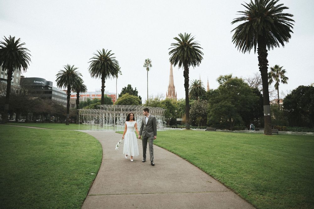 Couple walks hand in hand along a park path surrounded by palm trees and a church spire in the background.