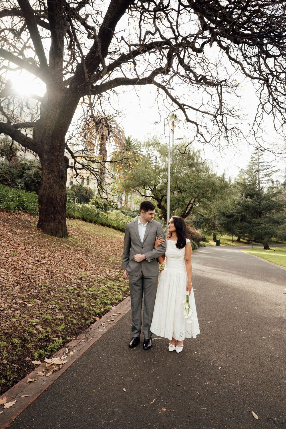 Bride and groom walk hand-in-hand on a park path, surrounded by bare trees and lush greenery on their wedding day.