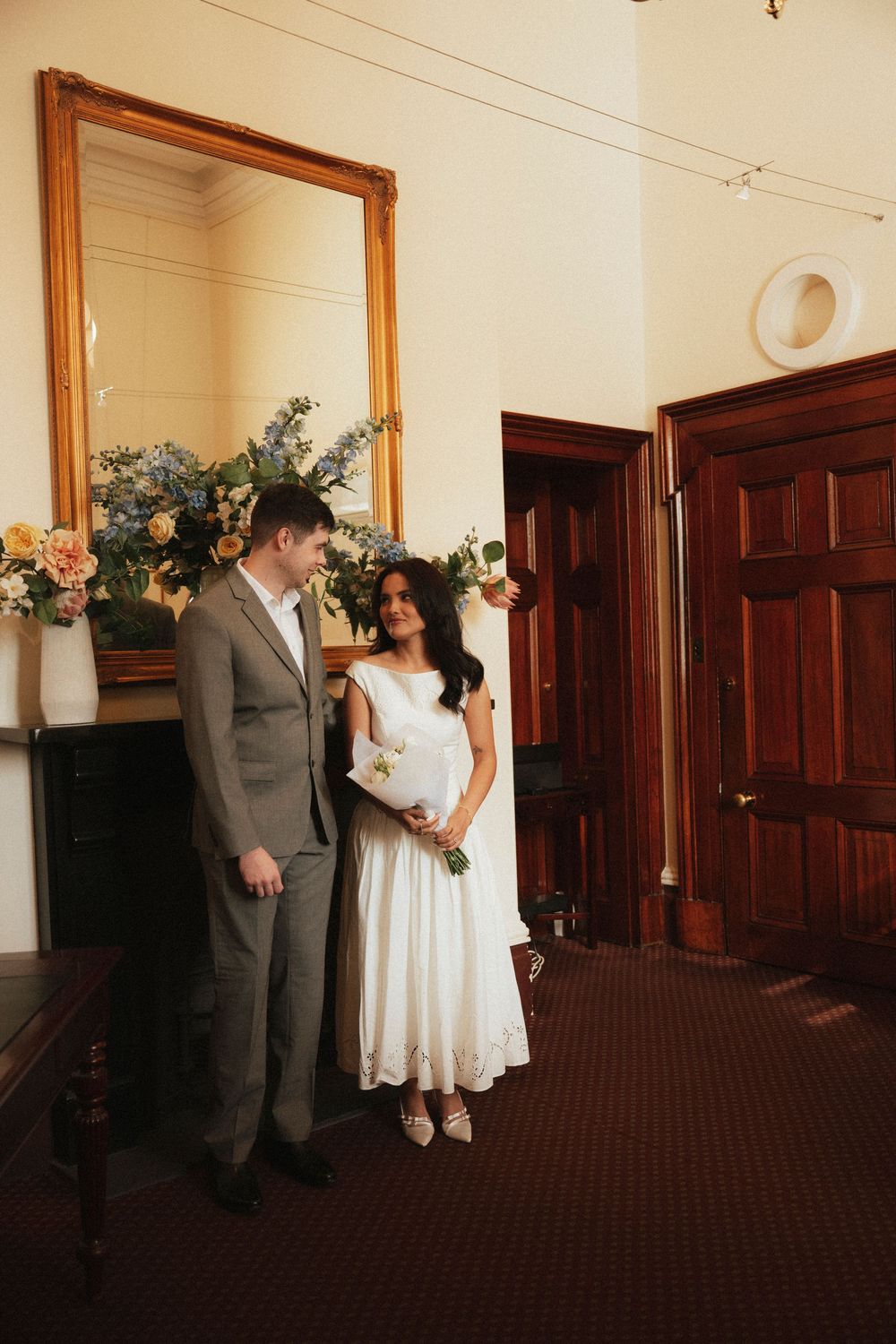 Bride in white midi dress and groom in gray suit stand together in elegant wedding venue with floral arrangements.